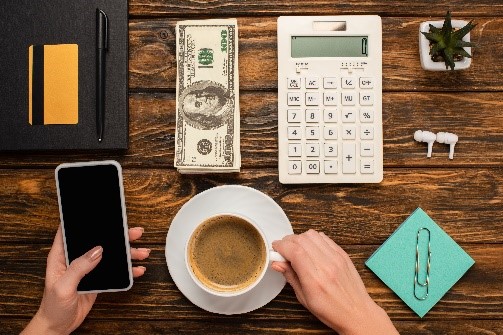 cropped view of a woman holding smartphone and coffee cup near dollar banknotes, calculator